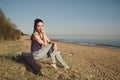 Pensive or thoughtful caucasian young woman in casual blue jeans, barefoot sitting on a wooden log the coast river Royalty Free Stock Photo