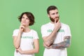 Pensive two young friends couple in white volunteer t-shirt isolated on green background. Voluntary free work assistance