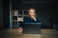 Pensive woman in a blue shirt sits in a cozy room at home and works on a laptop with a serious face. Female freelancer working Royalty Free Stock Photo