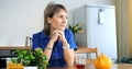 Pensive woman procrastinates at kitchen table, distant gaze. Conceptual procrastination, tea untouched, decision delayed