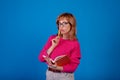 Pensive writer ponders new ideas, holds a red notepad and pencil near her mouth, has a pensive expression, wears big round glasses Royalty Free Stock Photo