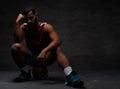 Pensive young African-American basketball player in sportswear sitting on a ball over dark background. Royalty Free Stock Photo