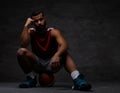 Pensive young African-American basketball player in sportswear sitting on a ball over dark background. Royalty Free Stock Photo