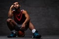 Pensive young African-American basketball player in sportswear sitting on a ball over dark background. Royalty Free Stock Photo