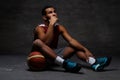 Pensive young African-American basketball player in sportswear sitting on a floor with a ball over dark background. Royalty Free Stock Photo