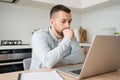 Pensive young Caucasian man sit at table at home kitchen work online on laptop pondering thinking. Thoughtful millennial Royalty Free Stock Photo