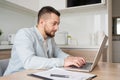 Pensive young Caucasian man sit at table at home kitchen work online on laptop pondering thinking. Thoughtful millennial Royalty Free Stock Photo