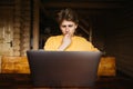 Pensive young freelancer man sitting at a table and working online on a laptop in a wooden country house Royalty Free Stock Photo