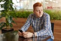 A pensive young handsome man with red hair and freckles sitting at the wooden table holding mobile phone in his hands looking asid Royalty Free Stock Photo