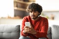 Pensive Young Indian Man Looking At Smartphone Screen While Sitting At Home Royalty Free Stock Photo