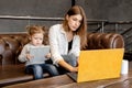 Pensive young mother sitting on sofa with little daughter, using laptop, working distantly at home. Attractive business Royalty Free Stock Photo