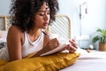 Pensive young multiracial latina woman lying down on bed with serious expression writing on journal in cozy bedroom Royalty Free Stock Photo