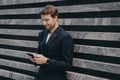 Pensive young office worker in formal stylish suit standing outdoor holding mobile phone in hands