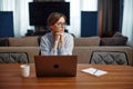 Pensive young woman in glasses sitting at desk with laptop looking aside, solving task, lost in thoughts, planning work
