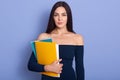 Pensive young woman holding paper folders in hands. Beautiful brunette woman wearing stylish dress standing isolated over blue Royalty Free Stock Photo