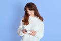 Pensive young woman wearing white casual knitted sweater using tobacco heating sticks looking on it isolated over blue background