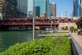 People cruise the riverwalk, boats cruise the river, el train crosses the river, and fishing poles rest on pier next to floating