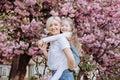 People, holidays and family concept - daughter hugs mom sitting on her back on cherry blossom background. Walk in the sakura trees