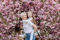 People, holidays and family concept - daughter hugs mom sitting on her back on cherry blossom background. Walk in the sakura trees