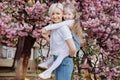 People, holidays and family concept - daughter hugs mom sitting on her back on cherry blossom background. Walk in the sakura trees