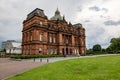 People Palace in Glasgow Green park which is used as a museum with people relaxing on a lawn with a white greenhouse in the back