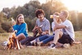 People, picnic and lifestyle concept. Joyful four friends sing song and play guitar, sit near bonfire, enjoy summer drink, breath