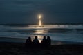 people sitting on the beach, watching a lighthouse beam light over the waves