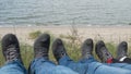 People sitting with legs outstretched on hill, three pairs of feet spread in gray sneakers on backdrop of grass and blue sea Royalty Free Stock Photo