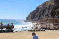 People sitting on a wooden benches overlooking the vast blue ocean water near a staircase covered with a rust fence and waves Royalty Free Stock Photo