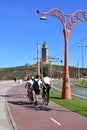 People skating and cycling in a promenade. Coastal public park with monument, grass, road, tram lines and street light. Sunny day.