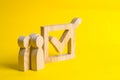 People stand near a wooden checkmark in a box on a yellow background. The concept of suffrage, voting in elections.