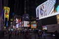 People stroll along Broadway in nighttime New York City amid glowing LED advertising panels on skyscrapers.