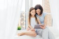 People, technology, family, children concept. Positive young other and her small daughter sit on window sill, hold digital tablet, Royalty Free Stock Photo