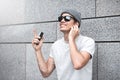 Cheerful guy dressed in gray t-shirt, sunglasses and hat at the street, listening to music with earphones, holding mobile phone Royalty Free Stock Photo