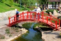 People walk across old red wooden bridge across a small river in green park. Vintage Japanese style bridge is reflected in the Royalty Free Stock Photo