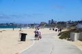 People walking along a smooth concrete bike trail along the beach with vast blue ocean water and buildings on the hillside