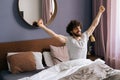 Peppy bearded young man waking up and stretching arms in morning sitting on bed. Happy handsome male have pleasant dream