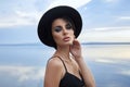 Perfect brunette beauty woman in a black hat and a black dress poses near a lake against a blue sky. Long hair woman and beautiful Royalty Free Stock Photo