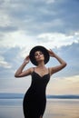 Perfect brunette beauty woman in a black hat and a black dress poses near a lake against a blue sky. Long hair woman and beautiful Royalty Free Stock Photo