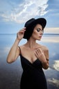 Perfect brunette beauty woman in a black hat and a black dress poses near a lake against a blue sky. Long hair woman and beautiful Royalty Free Stock Photo