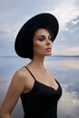 Perfect brunette beauty woman in a black hat and a black dress poses near a lake against a blue sky. Long hair woman and beautiful Royalty Free Stock Photo