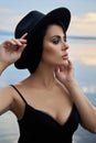 Perfect brunette beauty woman in a black hat and a black dress poses near a lake against a blue sky. Long hair woman and beautiful Royalty Free Stock Photo