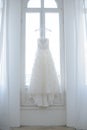 Perfect wedding dress with a full skirt and a train, sleeveless on a hanger in the bride`s room with white curtains. Royalty Free Stock Photo