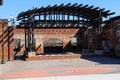 A performance stage made of red brick with a brown wooden pergola on top with tall black curved light posts i