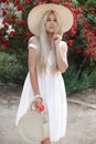 Young beautiful woman in a hat, near a large bush of red roses in the spring garden outdoors Royalty Free Stock Photo