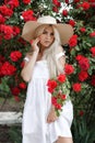 Young beautiful woman in a hat, near a large bush of red roses in the spring garden outdoors Royalty Free Stock Photo