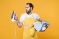 Perplexed young man househusband in apron hold basket with clean clothes, iron doing housework isolated on yellow