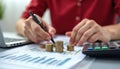 Person analyzes financial data on documents. Stacks of coins, calculator on table. Person uses pen, laptop. Office setting.