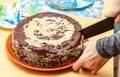 Person cutting, dividing a homemade chocolate cake decorated with many sprinkles using a knife, hand closeup. Home made cake