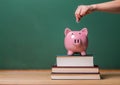 Person depositing money in a piggy bank on top of books with chalkboard Royalty Free Stock Photo